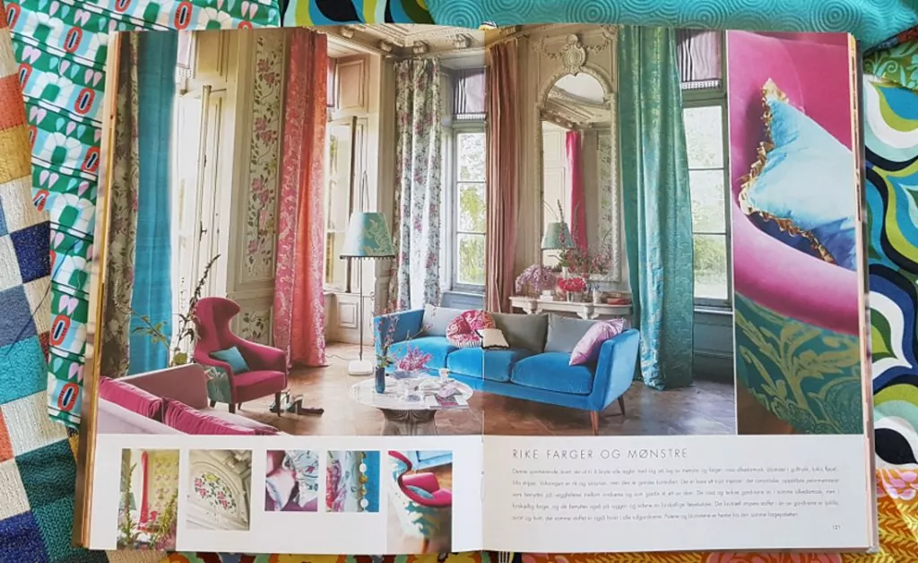 Tkaniny we wnętrzu,Album z fotografiami Tricia Guild leży na stole, widoczna fotografia salonu w stylu glamour, w tle kolorowe tkaniny