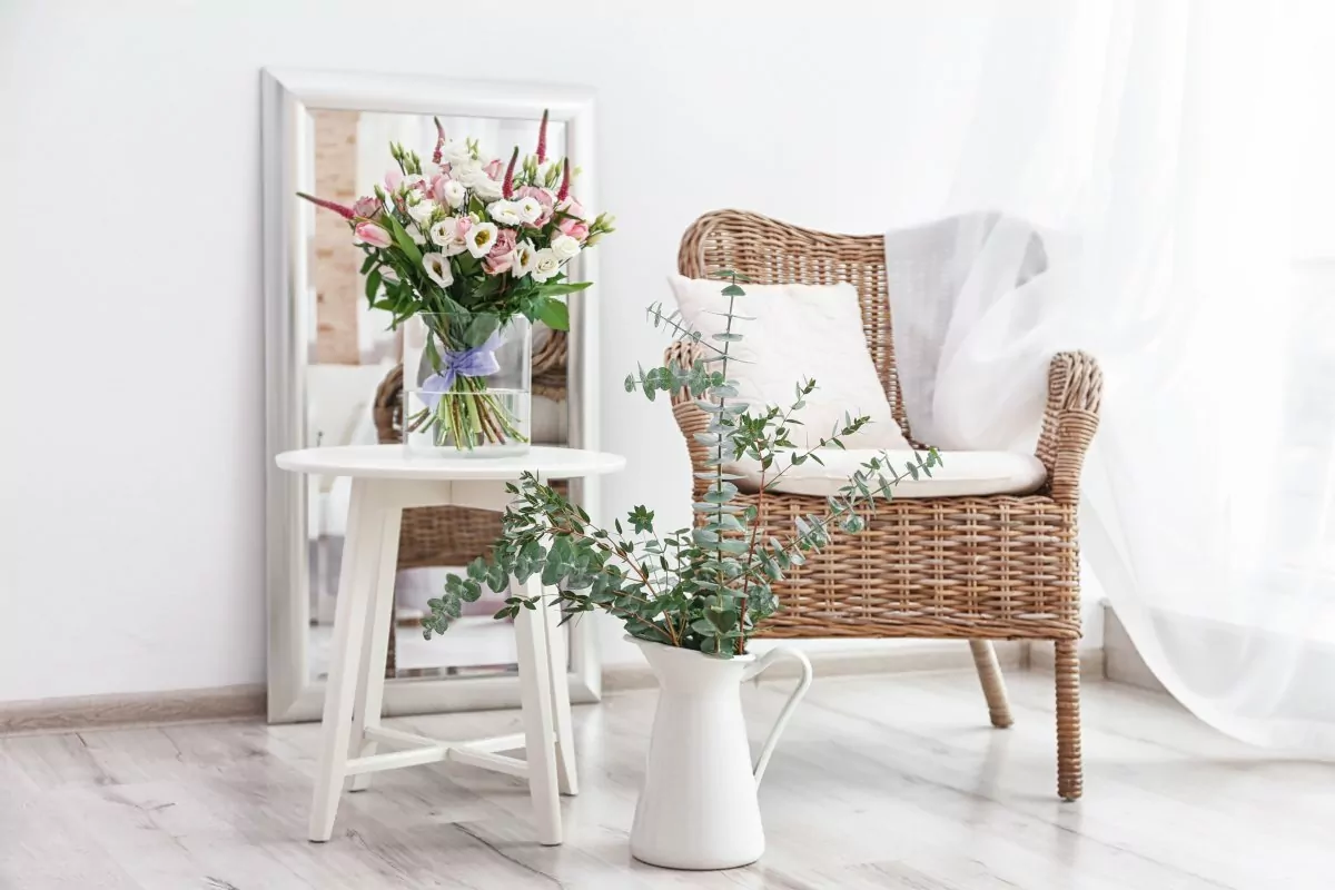 Dekoracje do salonu, zwiewna delikatka firanka, firanka przepuszczająca światło, fotel ratanowy z białą poduszką, biały stolik z wazonem i bukietem kwiatów, w tle stoi oparte o ścianę duże lustro w białej ramie
