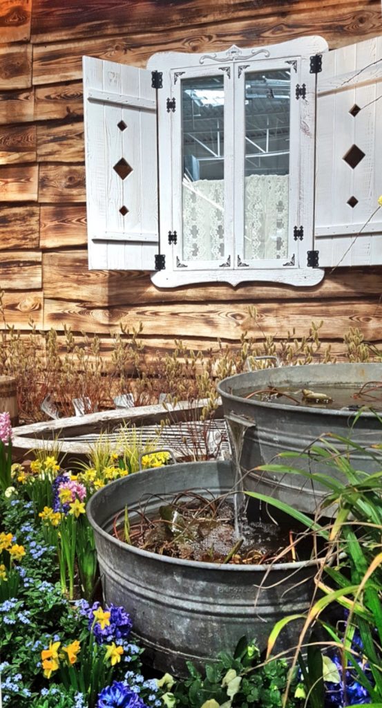 Targi Green Days, wystawa ogrodnicza, metalowe wanny, metalowa balia, źródełko ogrodowe, kolorowe kwiatki, żonkile, niezapominajki, w tle wiejska chata z białymi okiennicami