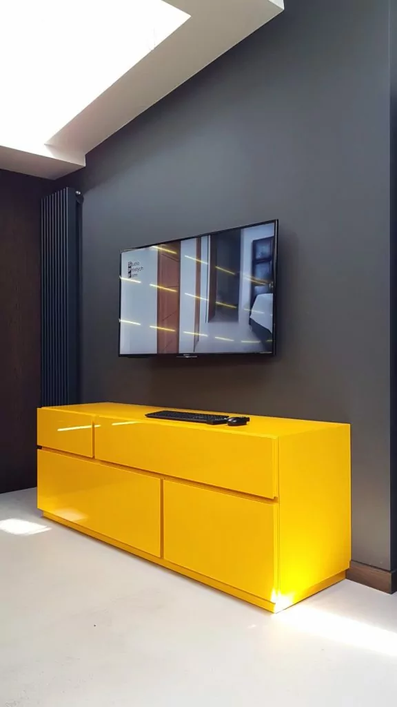 Ecomalta, Studio Prostych Form, żółta szafka RTV pod ścianą, na ścianie wisi telewizor, w rogu wysoki czarny grzejnik pionowy dekoracyjny, w tle szaara gładka ściana