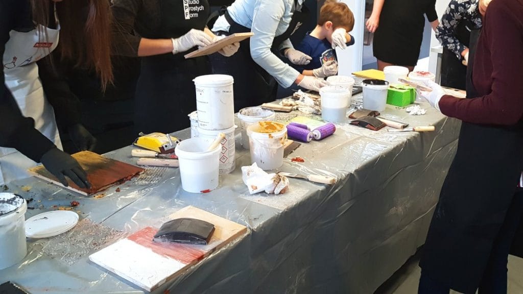 Ecomalta_warsztaty_1_1200_machinasnu.pl Ecomalta, okładzina, stół przygotowany do nakładania ecomalty, kilka opakowań pudełek wiaderek z kolorami ecomalty, deseczka z nałożoną ecomaltą, fioletowy wałek z odciskanym wzorem, wiele osób stoi przy stole i nakłada ecomalte na swoje deseczki, relacja z warsztatów ecomalty