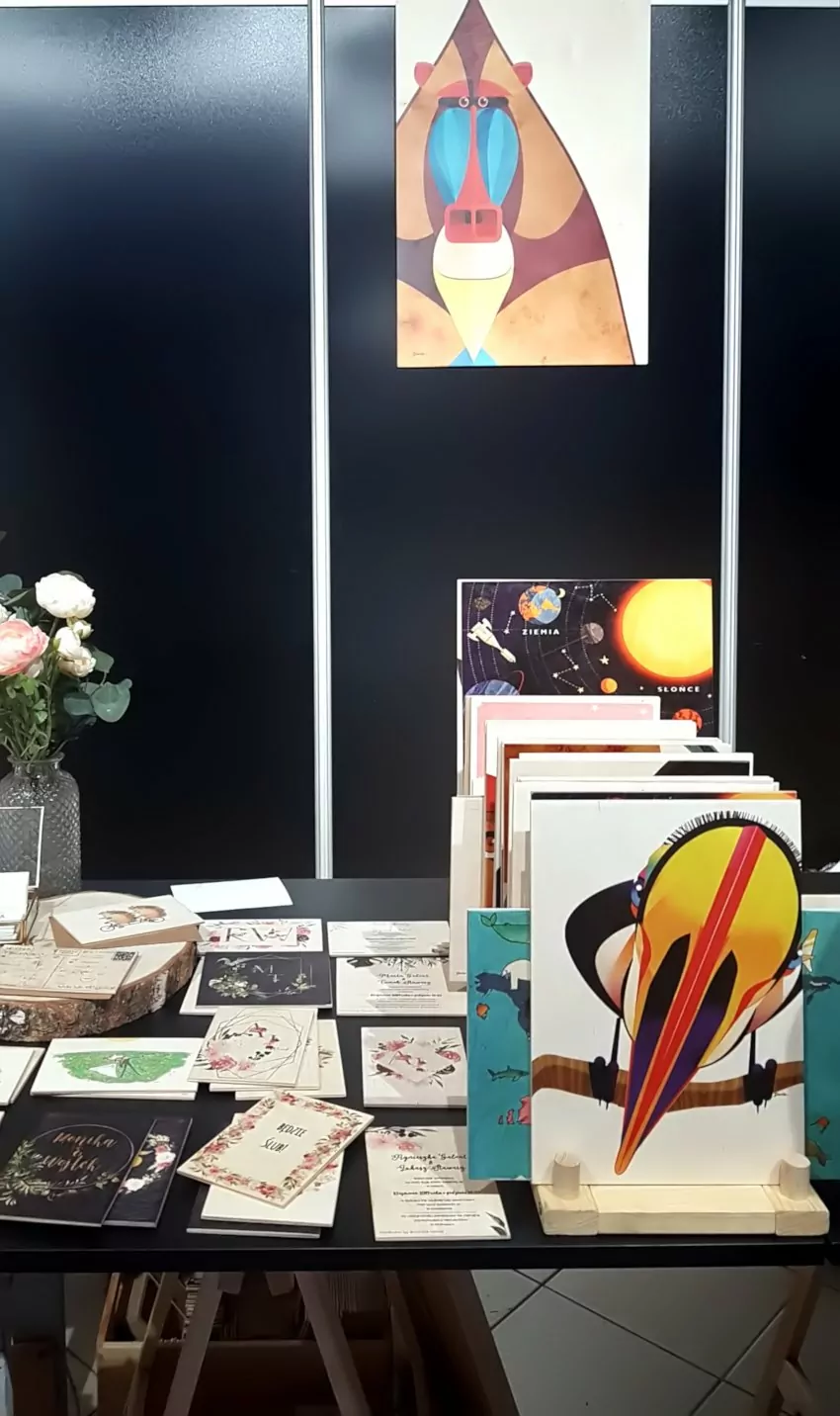 Targi Gift&Deco, drewniane deseczki leża na stole z czarnym obrusem, obok większe formaty są ustawione pionowo, na deseczkach kolorowe nadruki grafiki, drewniane kartki okolicznościowe z kolorowymi nadrukami
