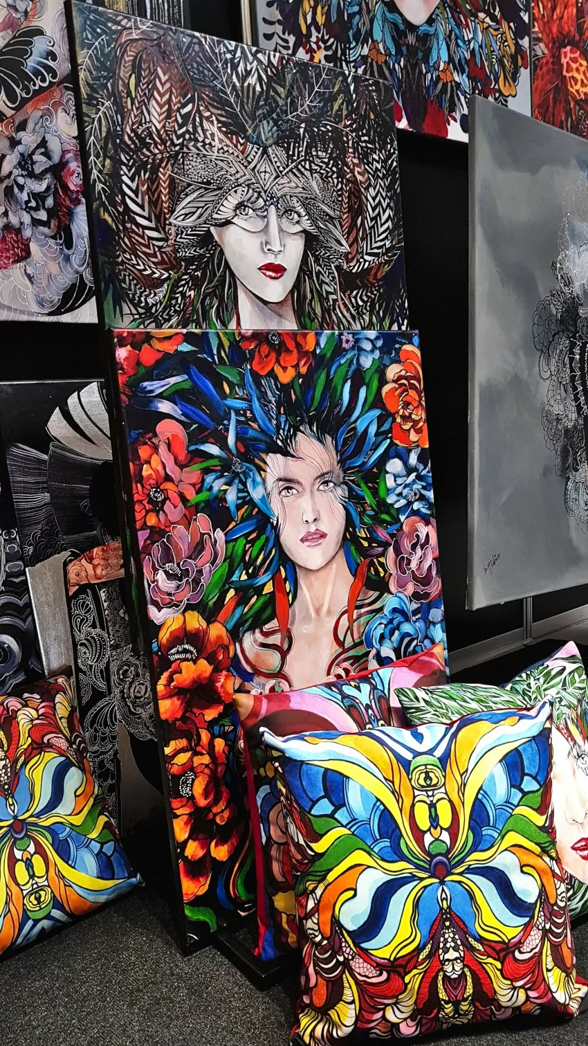 Targi Gift&Deco, obraz malarki Maggie Piu, obrazy z kobietami otoczone kolorowymi piórami i kwiatami, na podłodze położone kolorowe poduszki