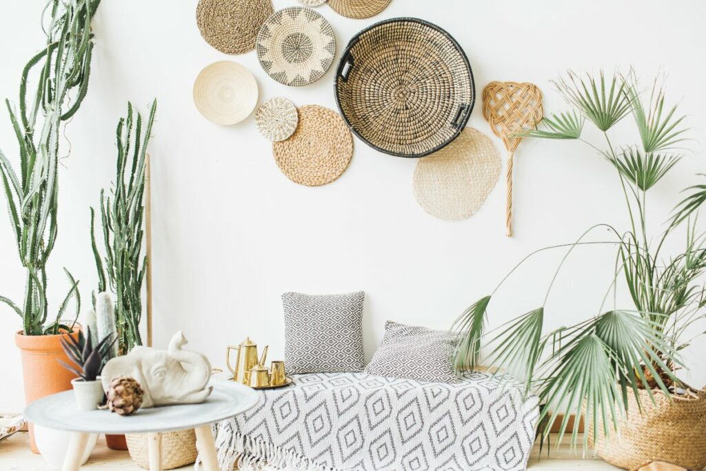 Dekoracje do salonu, na białej ścianie wiszą okrągłe plecionki z trawy morskiej, wikliny i sznurka, na drewnianej podłodzę stoją duże donice z roślinami, kaktusy w ceramicznych donicach, palma w wyklinowym koszyku