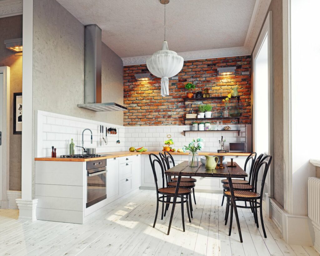 Kuchnia w stylu rustykalnym, na środku pomieszczenia stary prostokątny stół na cienkich nogach, krzesła z ratanowym siedziskiem, podłoga z bielonych desek, po lewej ciąg białych szafek kuchennych z drewnianym blatem, tle ściana z czerwonej cegły