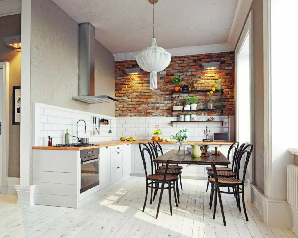 Kuchnia w stylu rustykalnym, na środku pomieszczenia stary prostokątny stół na cienkich nogach, krzesła z ratanowym siedziskiem, podłoga z bielonych desek, po lewej ciąg białych szafek kuchennych z drewnianym blatem, tle ściana z czerwonej cegły