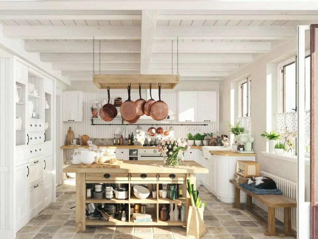 Kuchnia w stylu rustykalnym, na środku pomieszczenia drewniana wyspa pomocnik, nad nią wiszą patelnie po prawej pod oknem drewniana ławka na której śpi kot, po lewej biały rustykalny kredens w tle szafki kuchenne na ścianę z białych desek