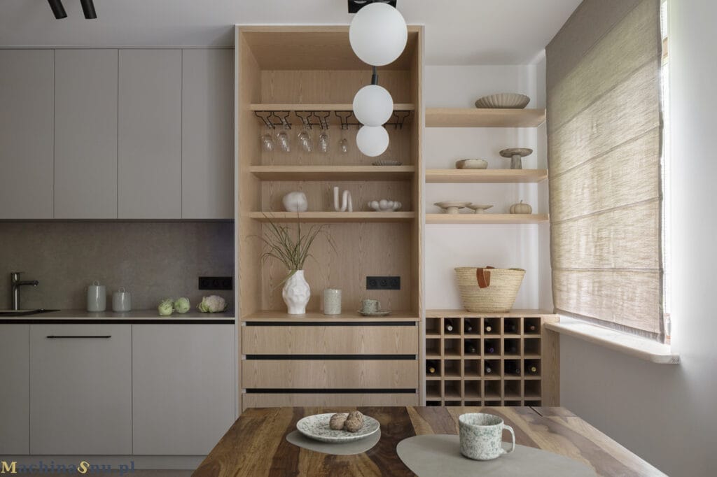 Wysoki, otwarty regał z jasnego dębu stojący przy ciemnym, drewnianym stole. Regał posiada p&oacute;łki, szuflady i winiarkę.