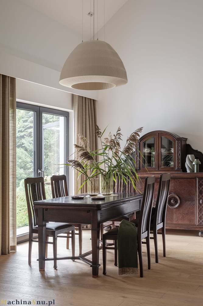 Antyczny, ciemnobrązowy stół z krzesłami i kredens stojące na jasnej, dębowej podłodze. Nad stołem wisi duża, nowoczesna lampa.