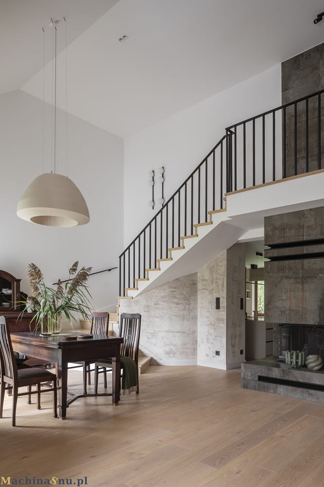 Wysoki salon z nowoczesnym kominkiem wykończonym ciemnym spiekiem o rdzawym wzorze. W tle widać antresolę z czarną, minimalistyczną balustradą i schody.