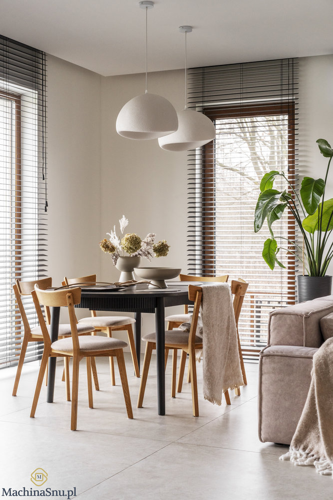 Czarny okrągły stół w jadalni, drewniane krzesła z jasną tapicerką i dwie białe, organiczne lampy wiszące. Duże okna z żaluzjami.
