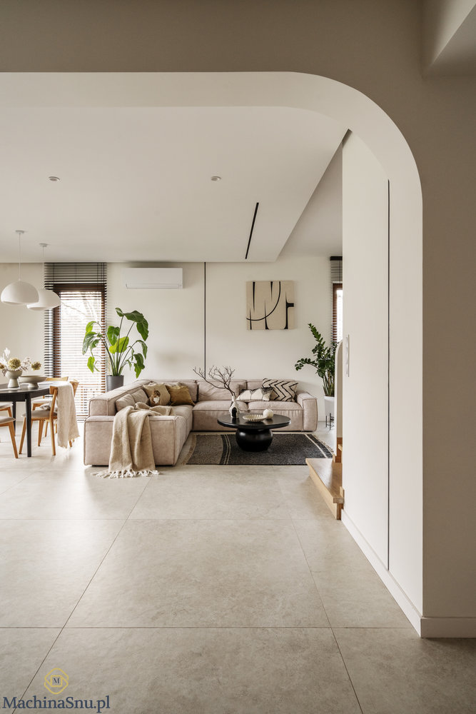 Widok ogólny na salon i jadalnię. Duża beżowa sofa narożna, czarny stół z krzesłami i białe lampy wiszące.