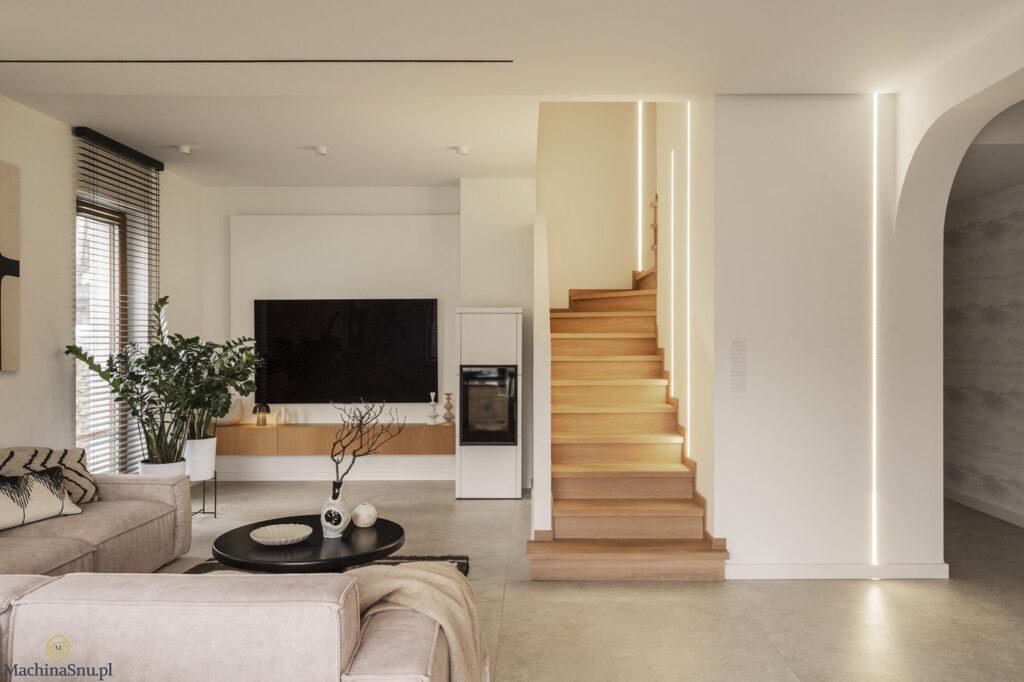Nowoczesny salon ze schodami dębowymi i pionowym oświetleniem LED w ścianie. Biały kominek Romotop i płytki Ground Arena.