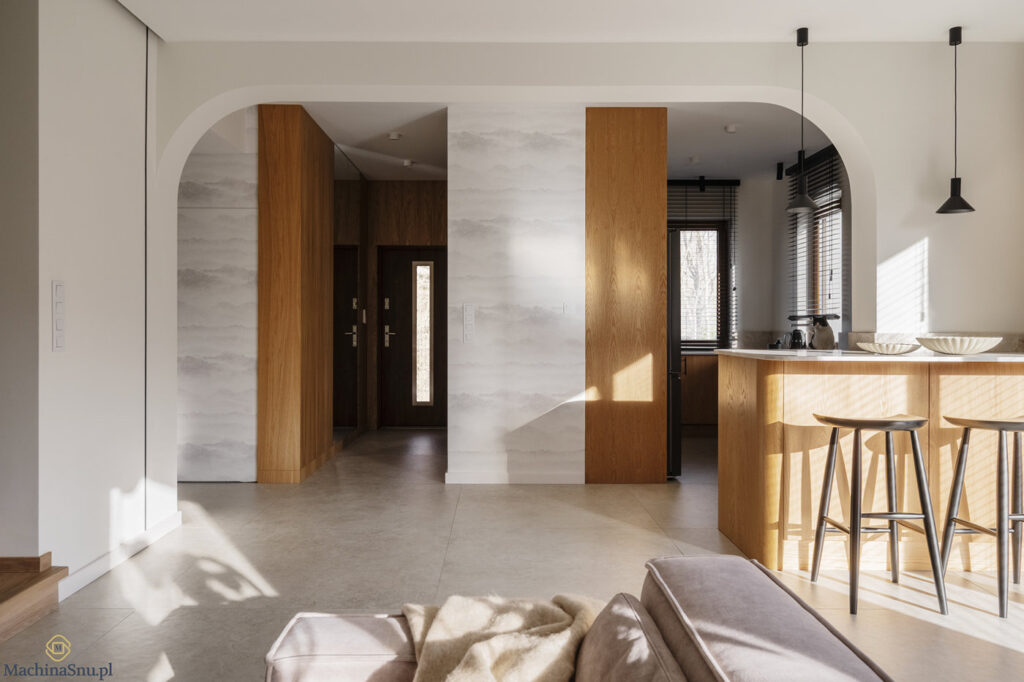 Widok og&oacute;lny na strefę dzienną: salon, kuchnię i korytarz. Dwa dekoracyjne łuki, tapeta z motywem lasu Rebel Walls i płytki wielkoformatowe.